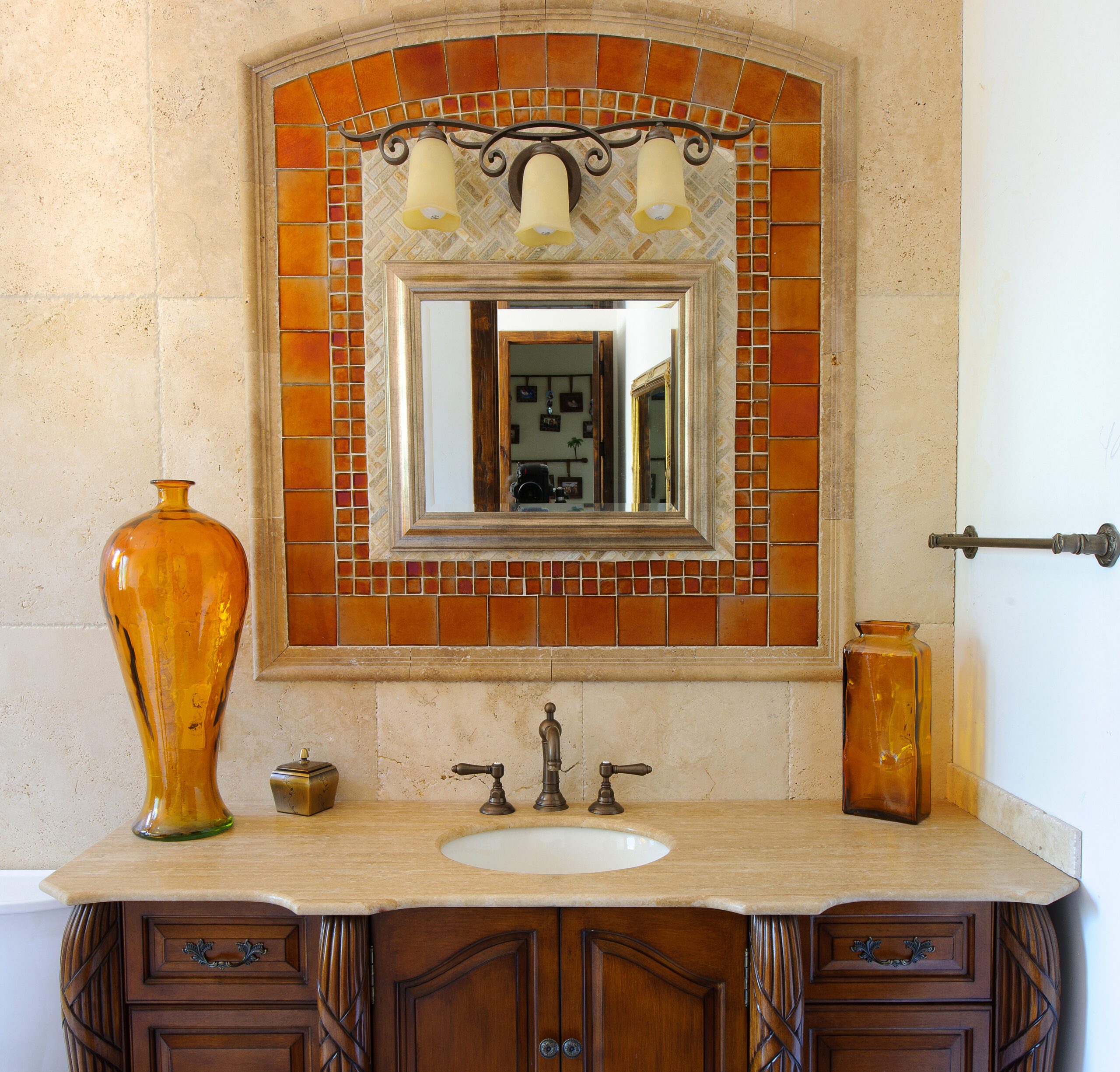 A traditional bathroom vanity with wooden cabinets and a stone countertop, featuring an orange tiled mirror and decorative items.
